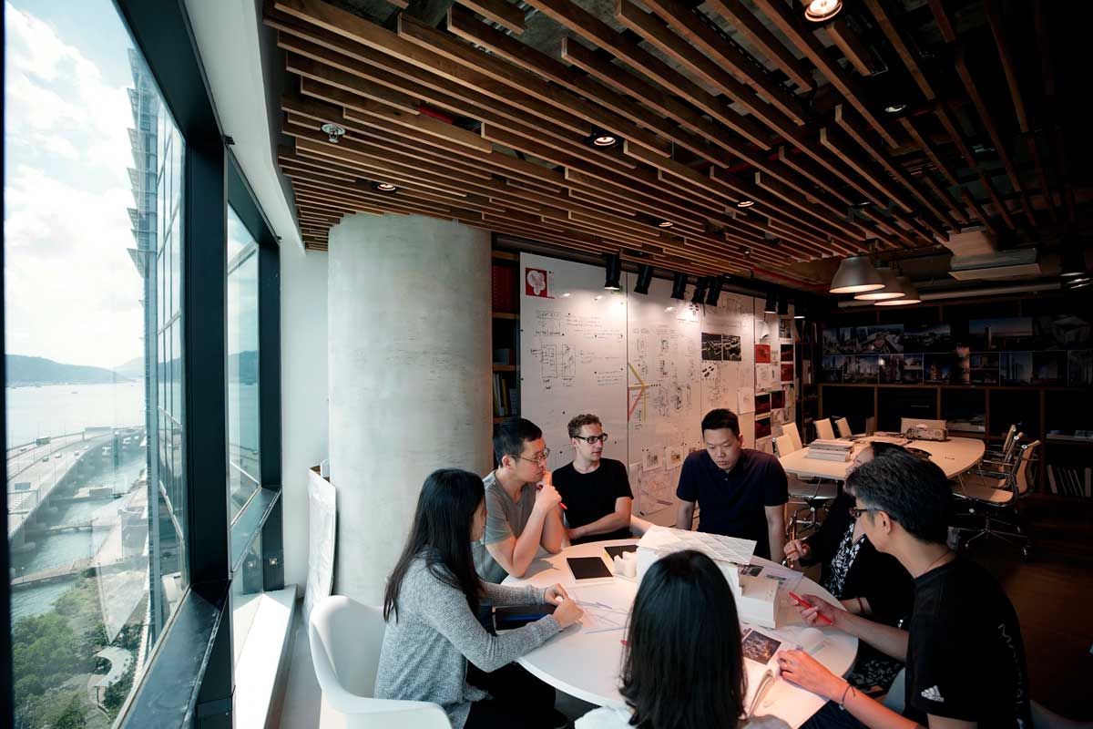 Group of people sat around table in the corner of a room. Lots of paper on the table.