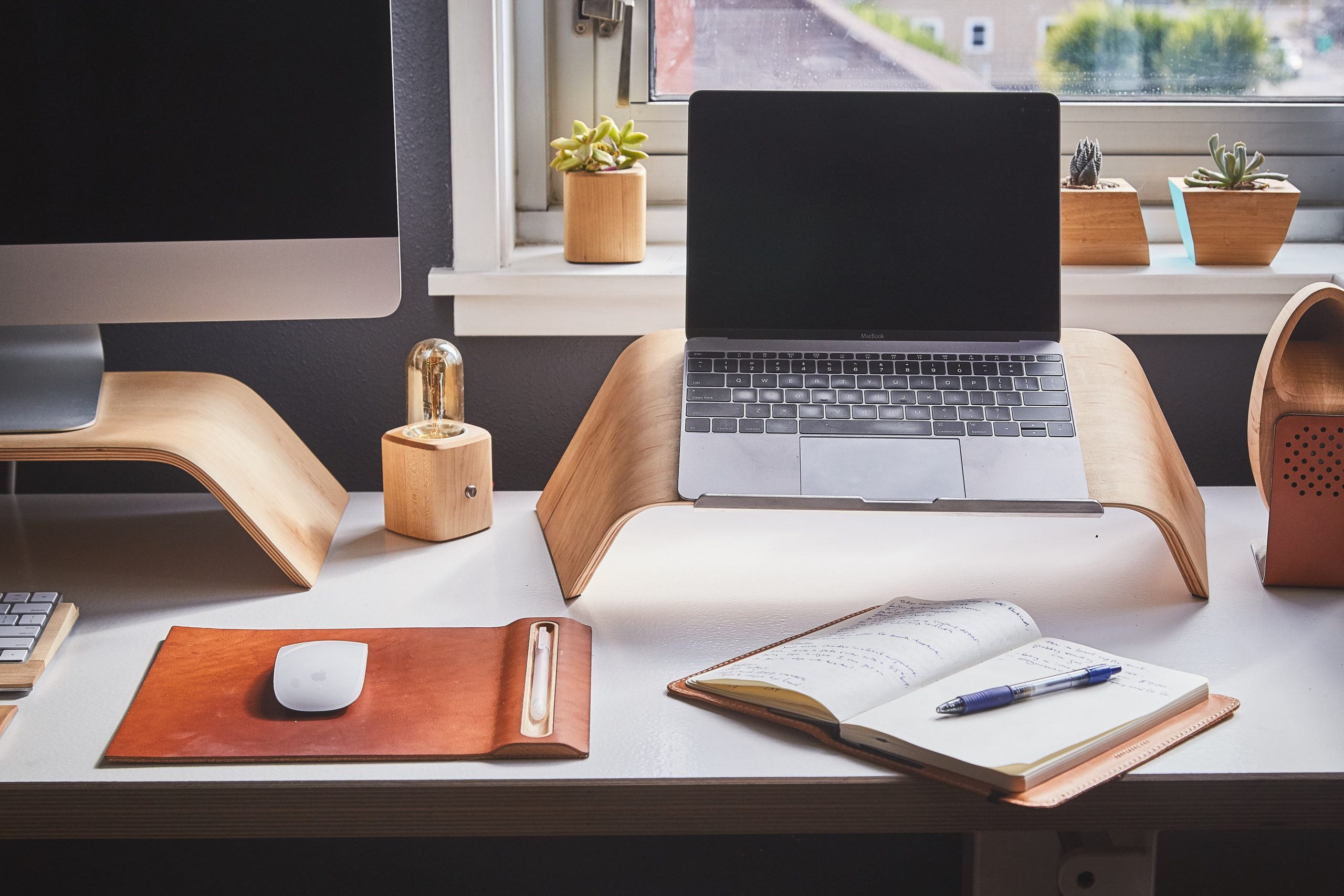 A laptop and computer monitor on a desk by a window with a notebook on the desk