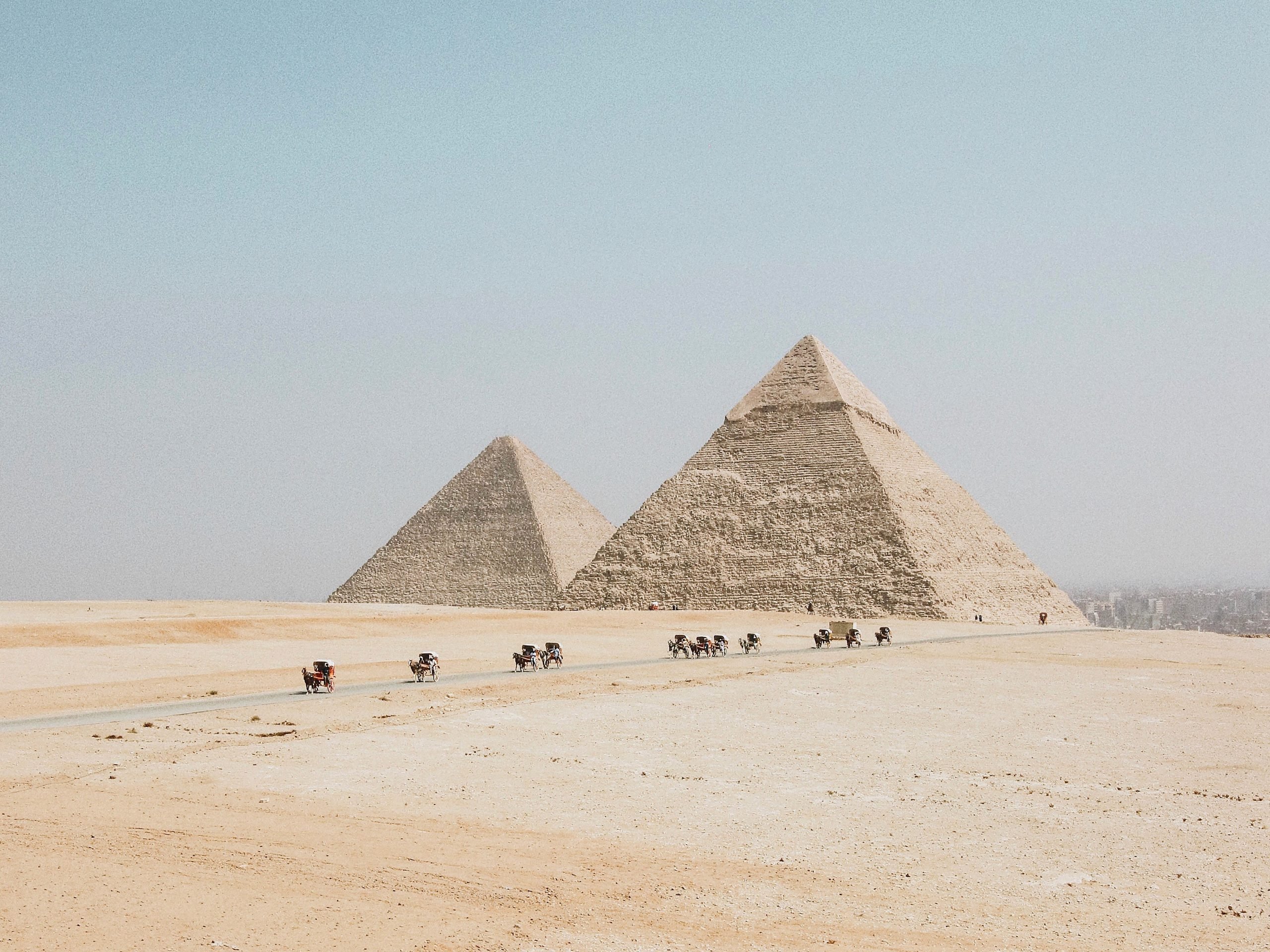 stone pyramids in desert, people and camels traveling on dirt road below